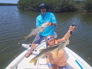 Two people on a boat, smiling and holding up fish. Setting is a waterway with mangroves.
