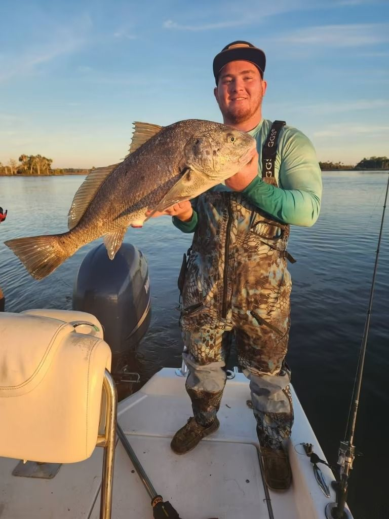 Man smiling, holding a reddish-brown fish on a boat, blue sky and mangroves in background.
