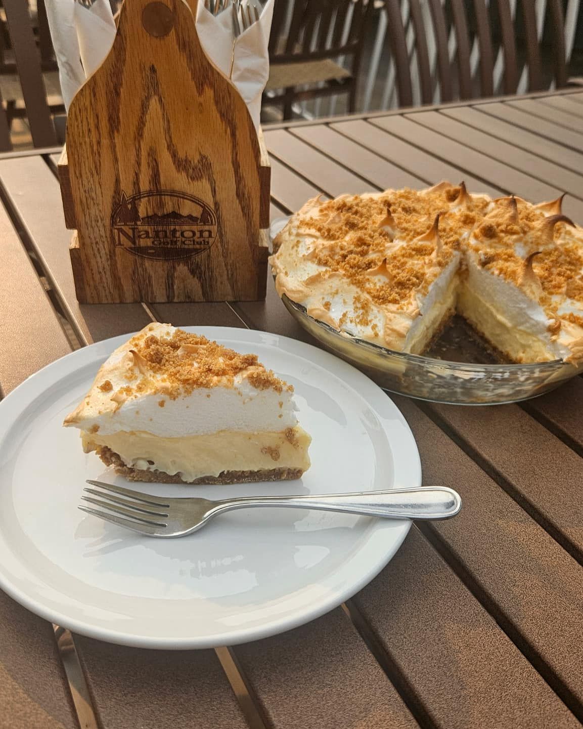 Slice of cream pie on a white plate beside a whole pie on a wooden table outdoors