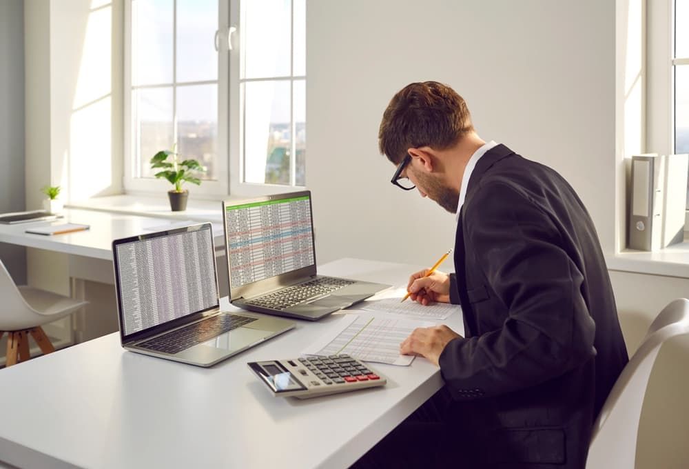 Man in Suit Working at A Desk Reviewing Papers While Using Two Laptops — Careful Books in Westcourt, QLD