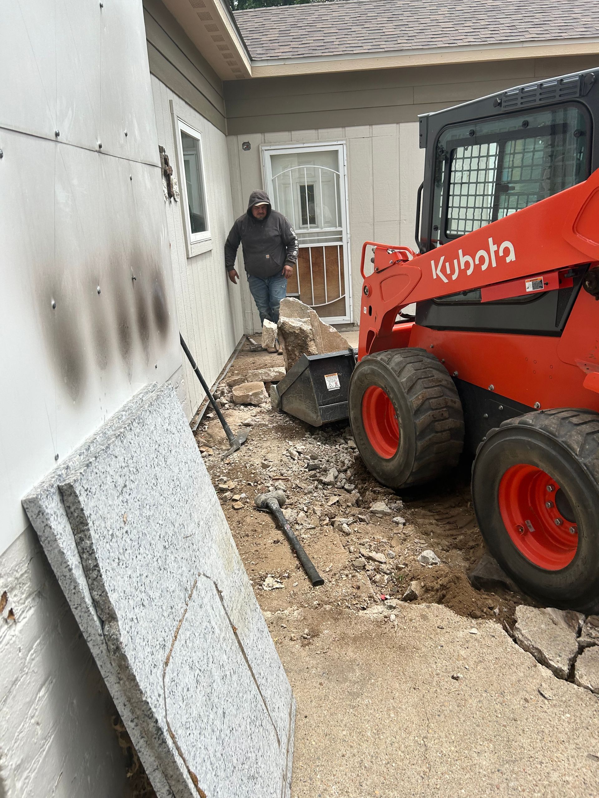 Worker beside a red Kubota skid steer clearing debris along a house exterior with gravel and broken concrete.