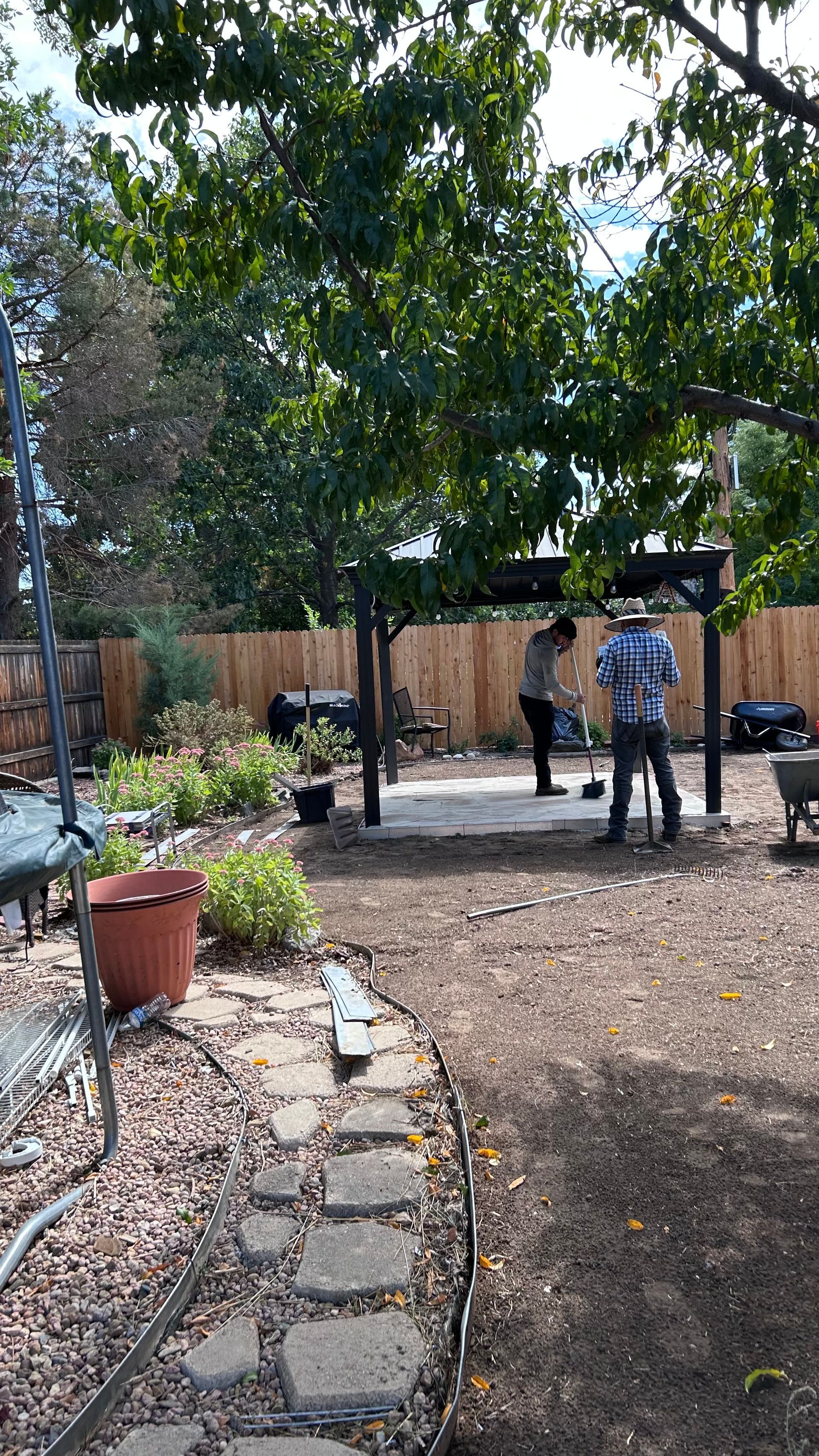 Backyard with trees, stone path, potted plants, and two people near a wooden fence.