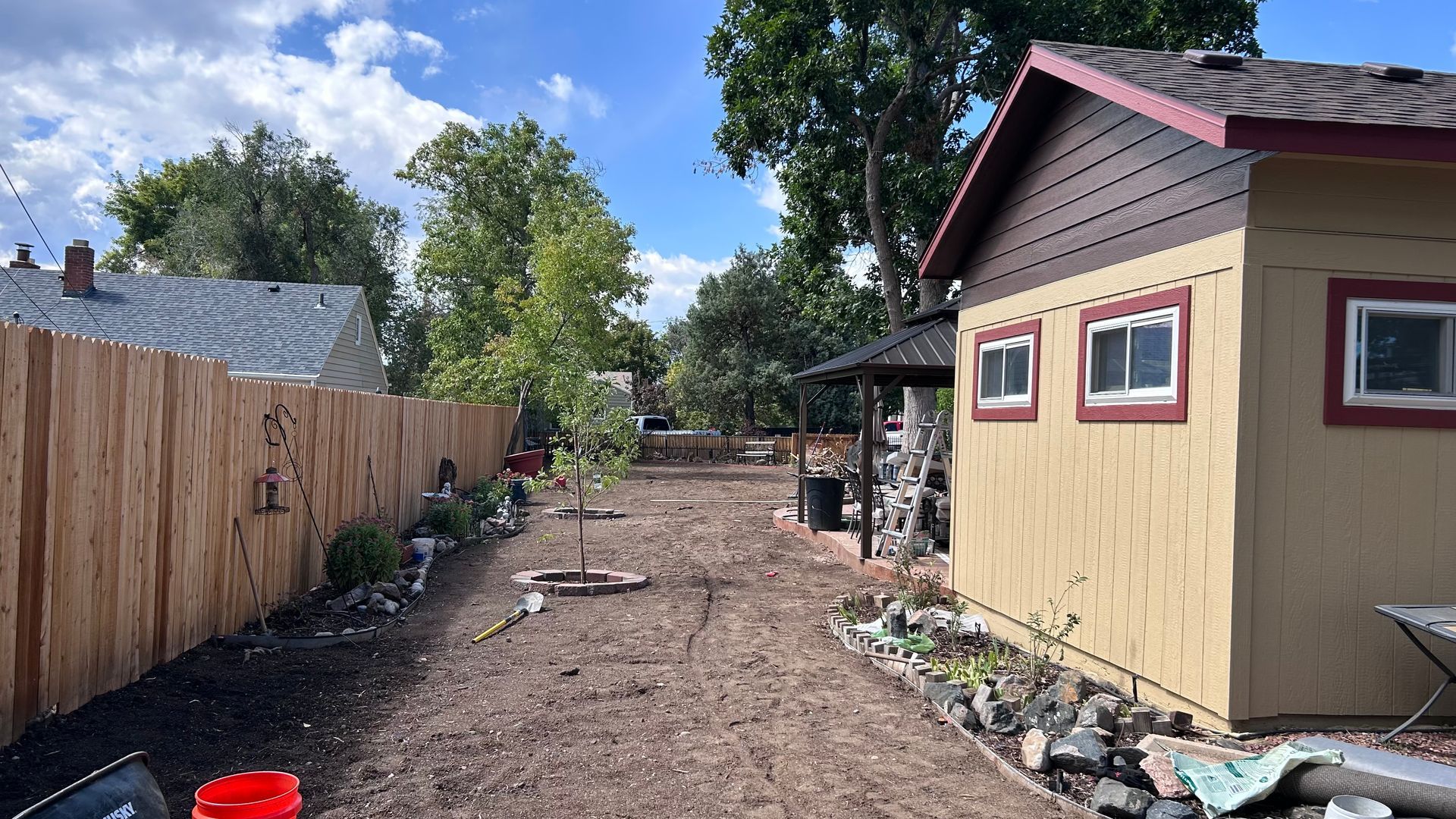 Backyard with wooden fence, tan shed, and dirt path under trees on a sunny day