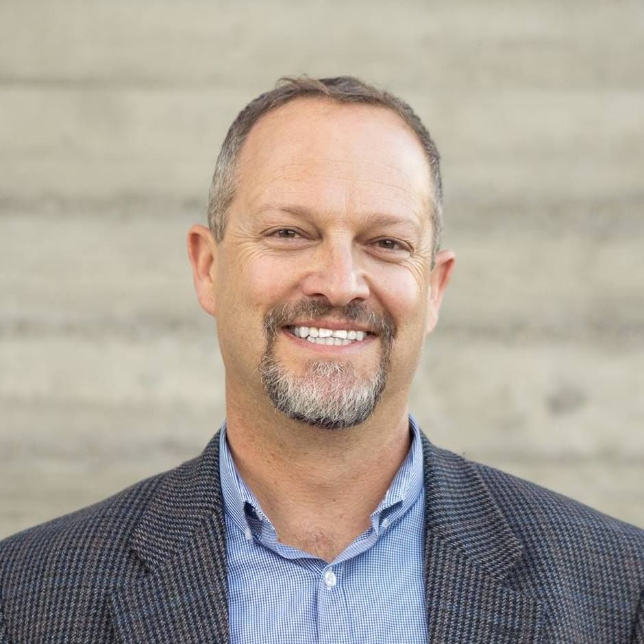 Man with short brown hair and goatee smiling, wearing a blue shirt and patterned blazer in front of a concrete wall.