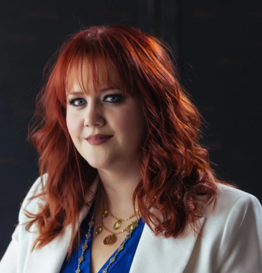 Woman with red wavy hair and bangs, wearing a white blazer and blue top, smiling.