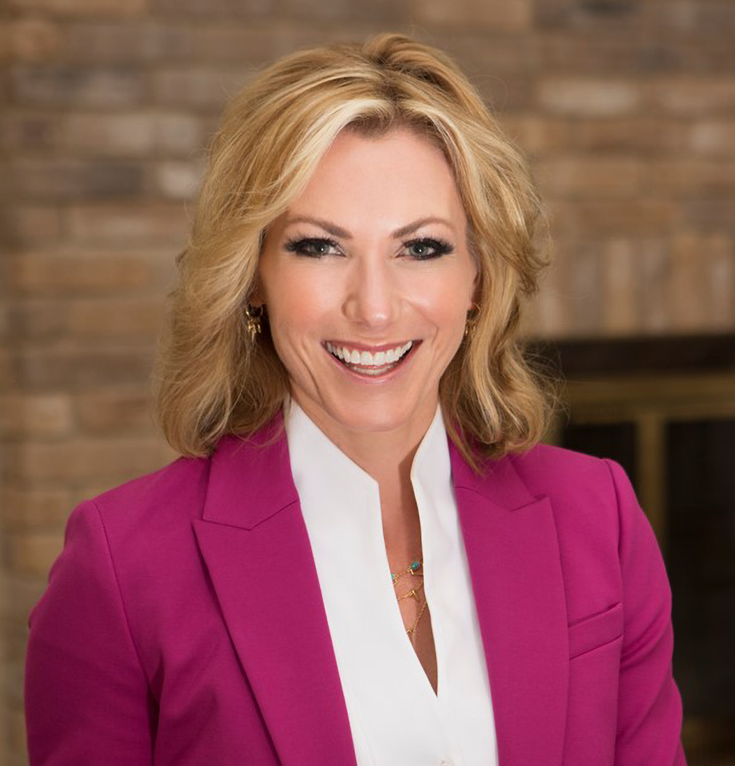 Woman smiling, wearing a pink blazer and white shirt, in front of a brick wall.