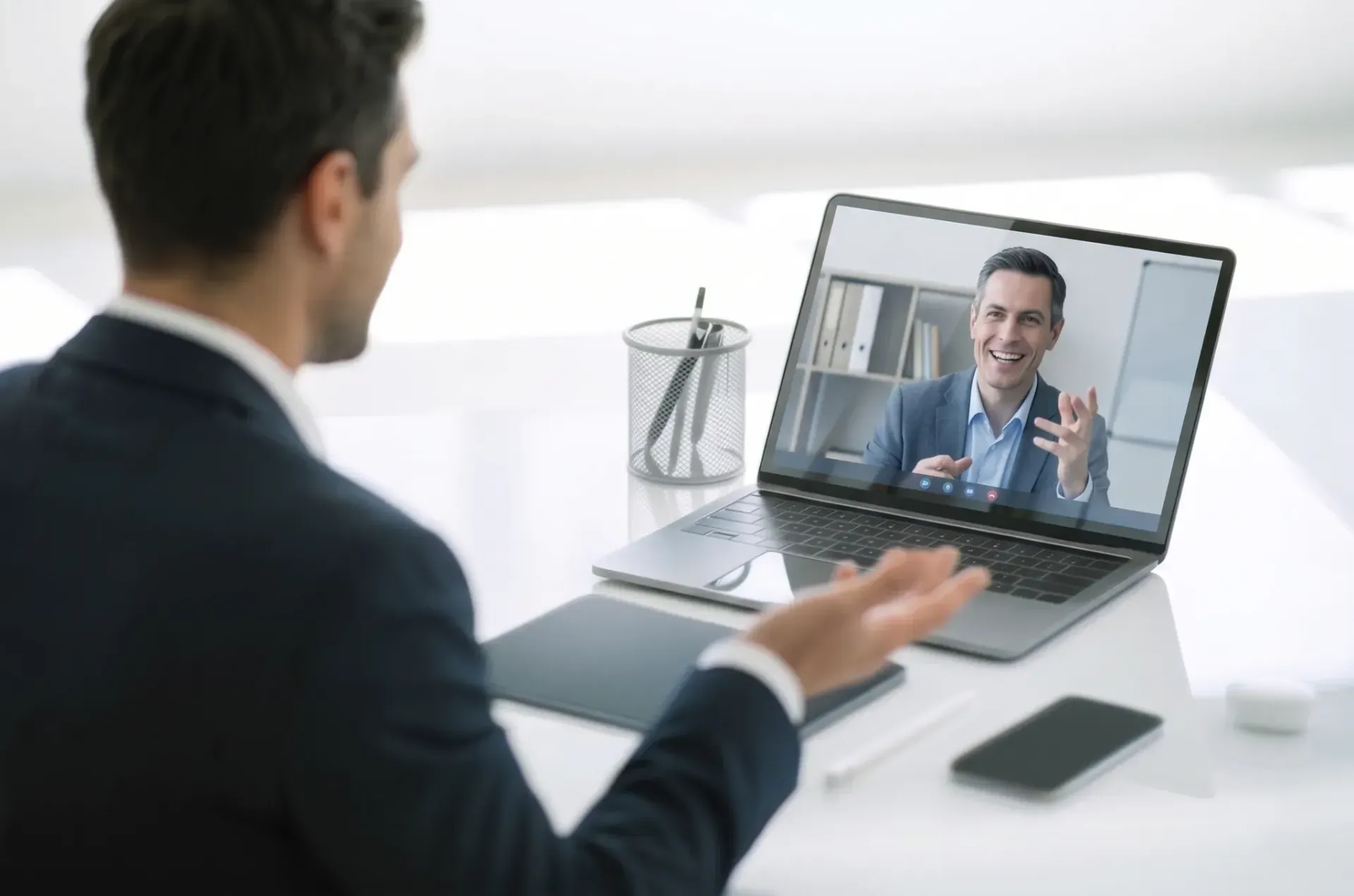 Man on a video call, gesturing toward a laptop screen in a bright office desk setup.