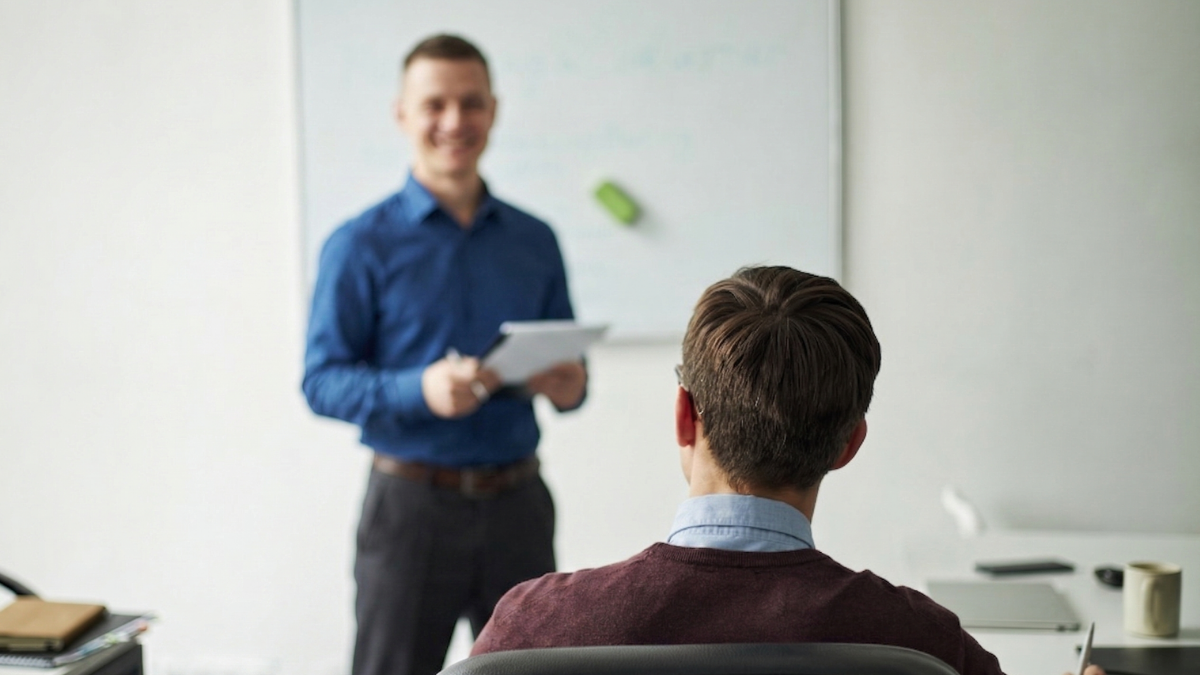 Presenter speaking to an audience in a classroom, holding papers beside a whiteboard.