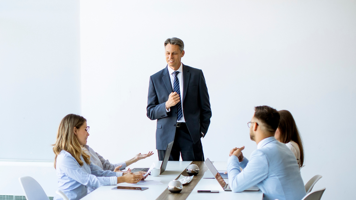 Business meeting in a bright conference room, with one man standing and speaking to three seated colleagues.