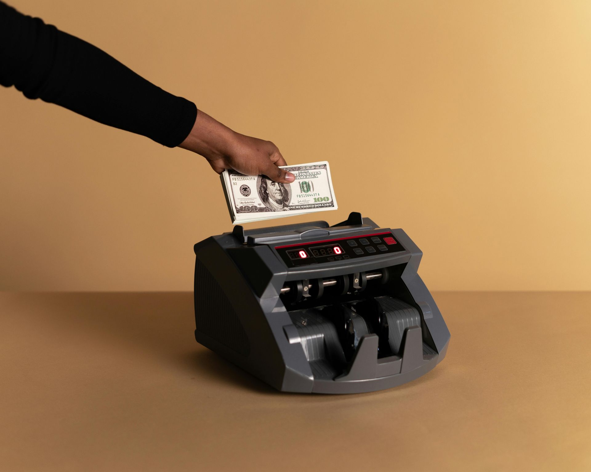 Hand inserts a dollar bill into a black money counter on a tan background.