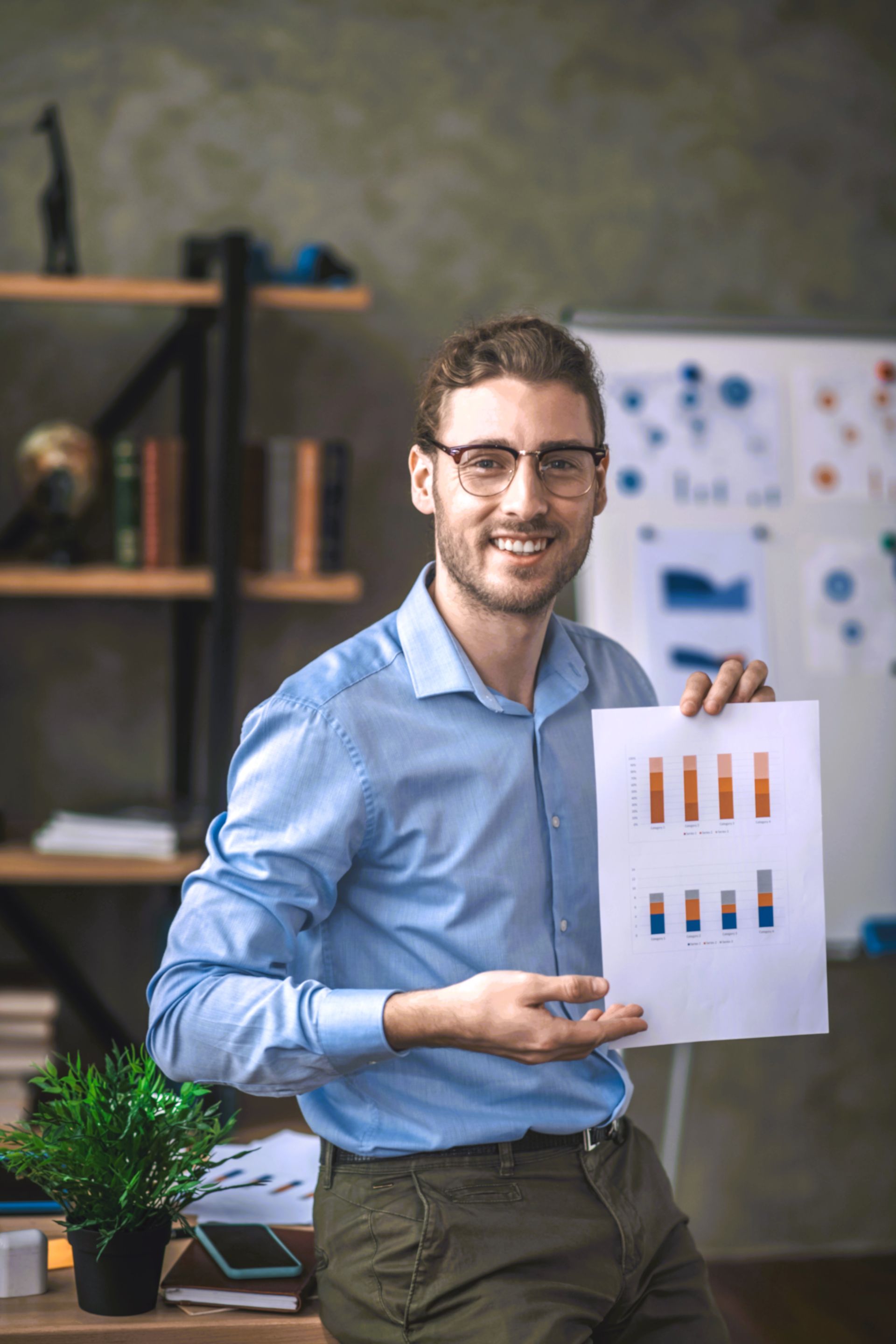 Man holding a paper with bar graphs, smiling. In an office setting, with a whiteboard and shelves in background.