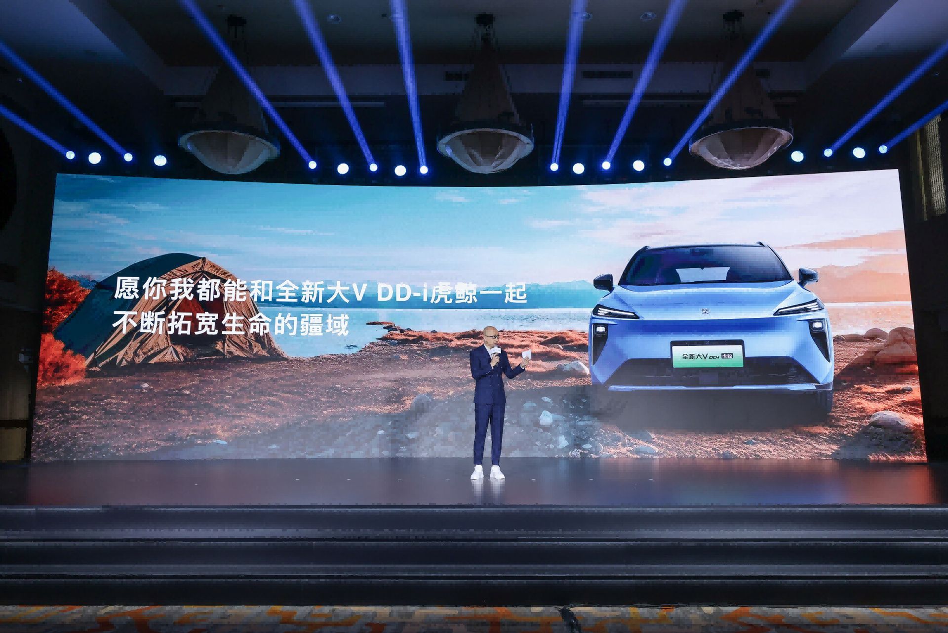 A presenter on stage gestures towards a large screen displaying a blue car and text in front of a natural backdrop.