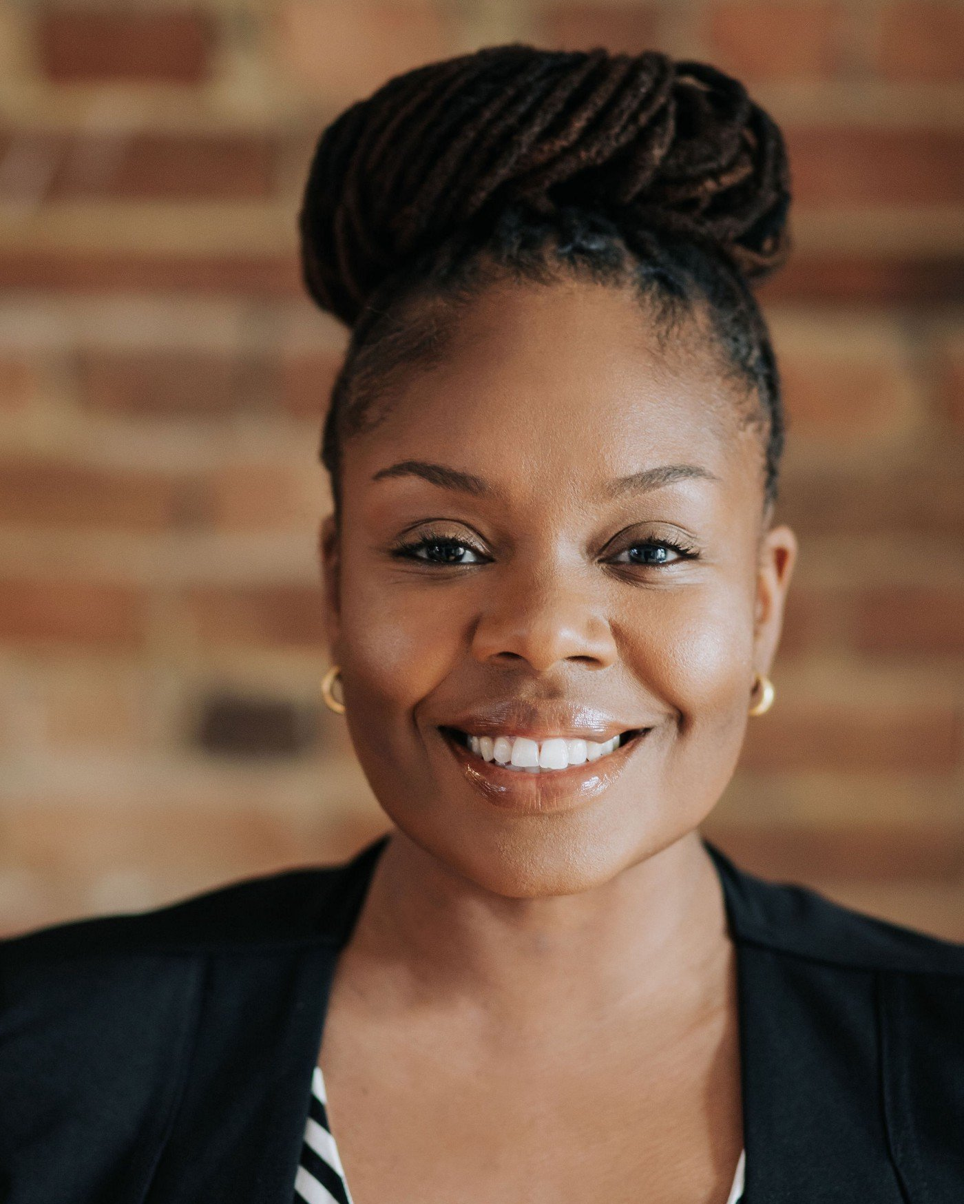 A woman with dreadlocks is smiling in front of a brick wall.