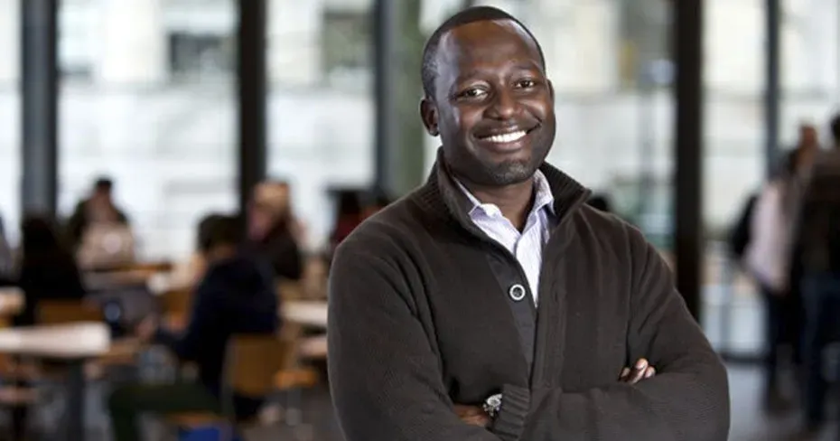 A man in a brown sweater is standing with his arms crossed and smiling.