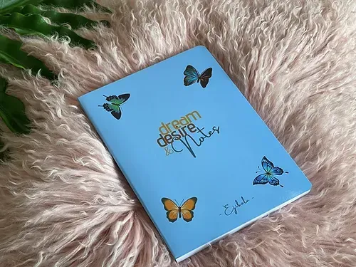 A blue notebook with butterflies on it is sitting on a pink blanket.