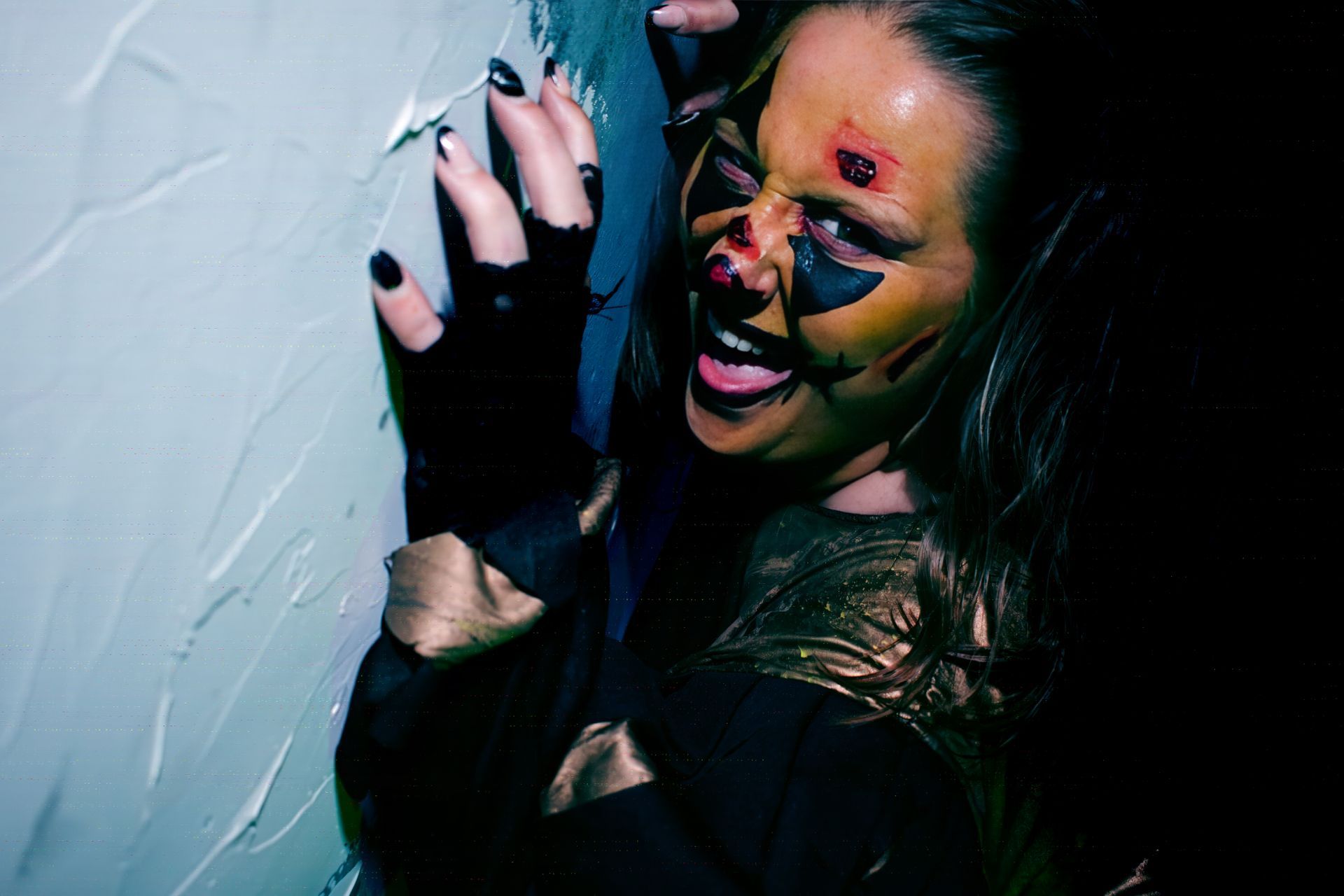 Person in black glove and shirt, face splattered with color, pressing against a pale wall with a grimacing expression