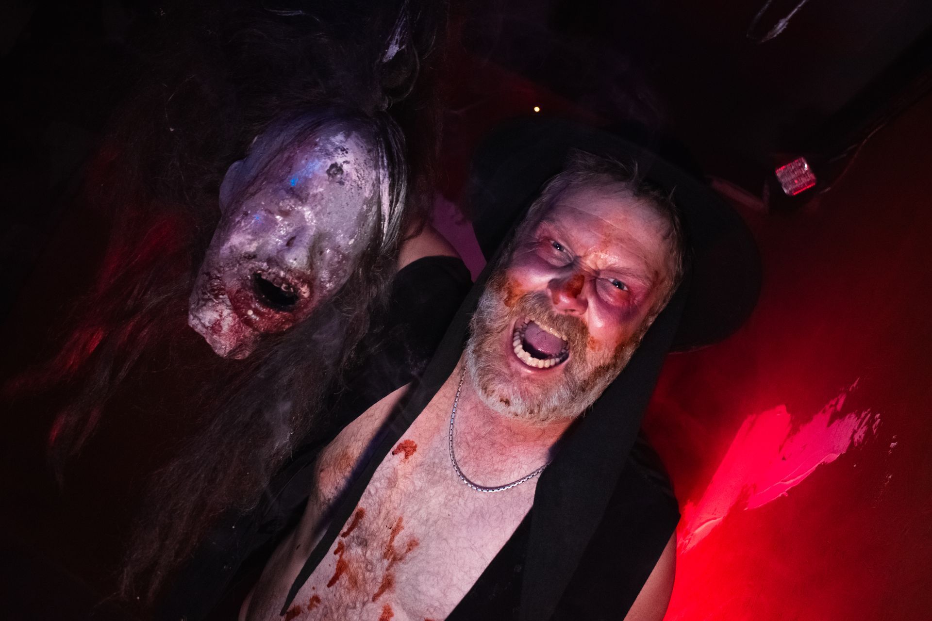 Two men in dark costumes, one with a pale mask, in a red-lit horror scene