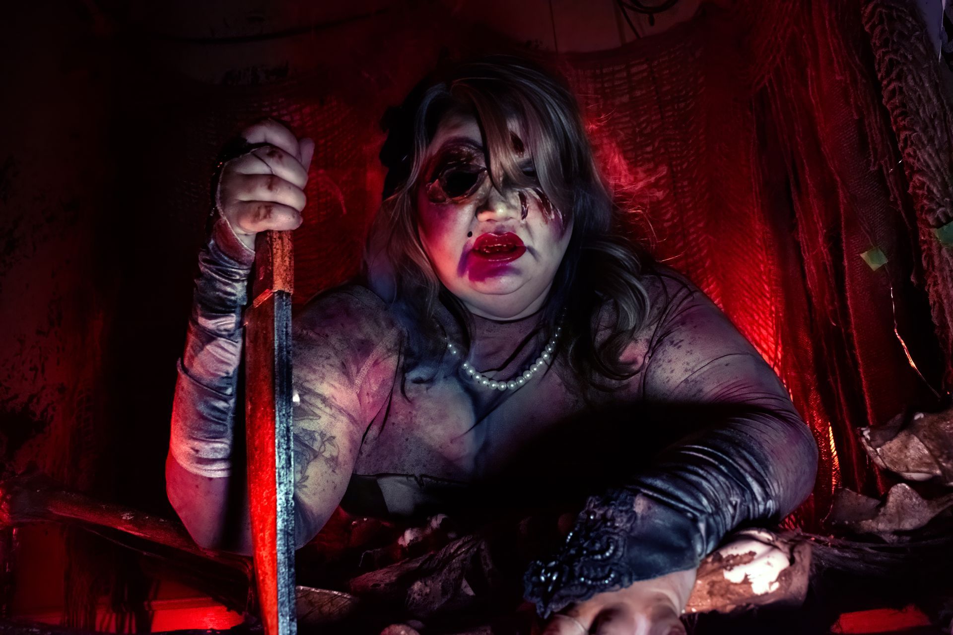 Gothic portrait of a person with dark makeup, holding a staff in red lighting and draped in black fabric