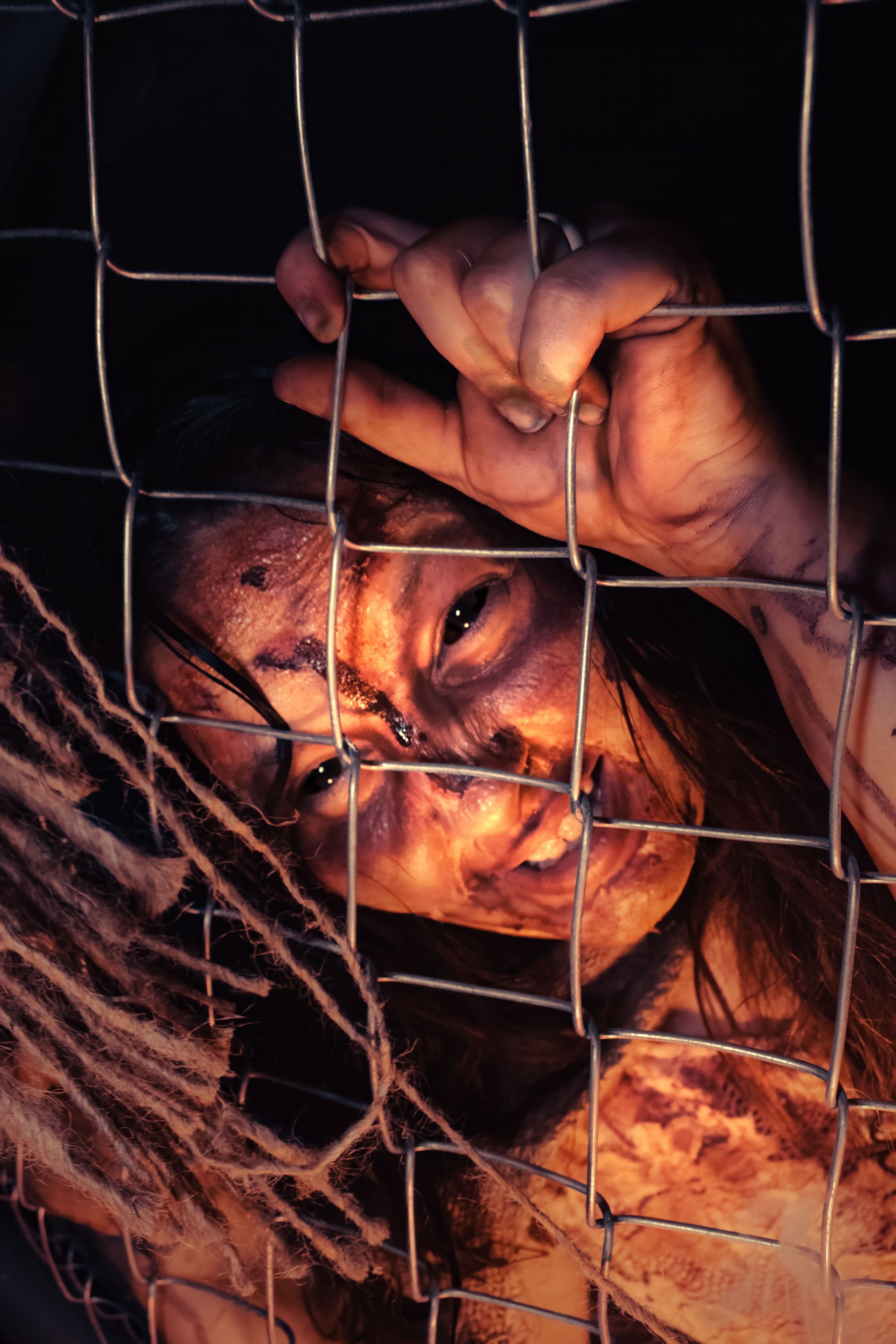 Person gripping a wire fence, face pressed close, with an intense expression and dim orange lighting