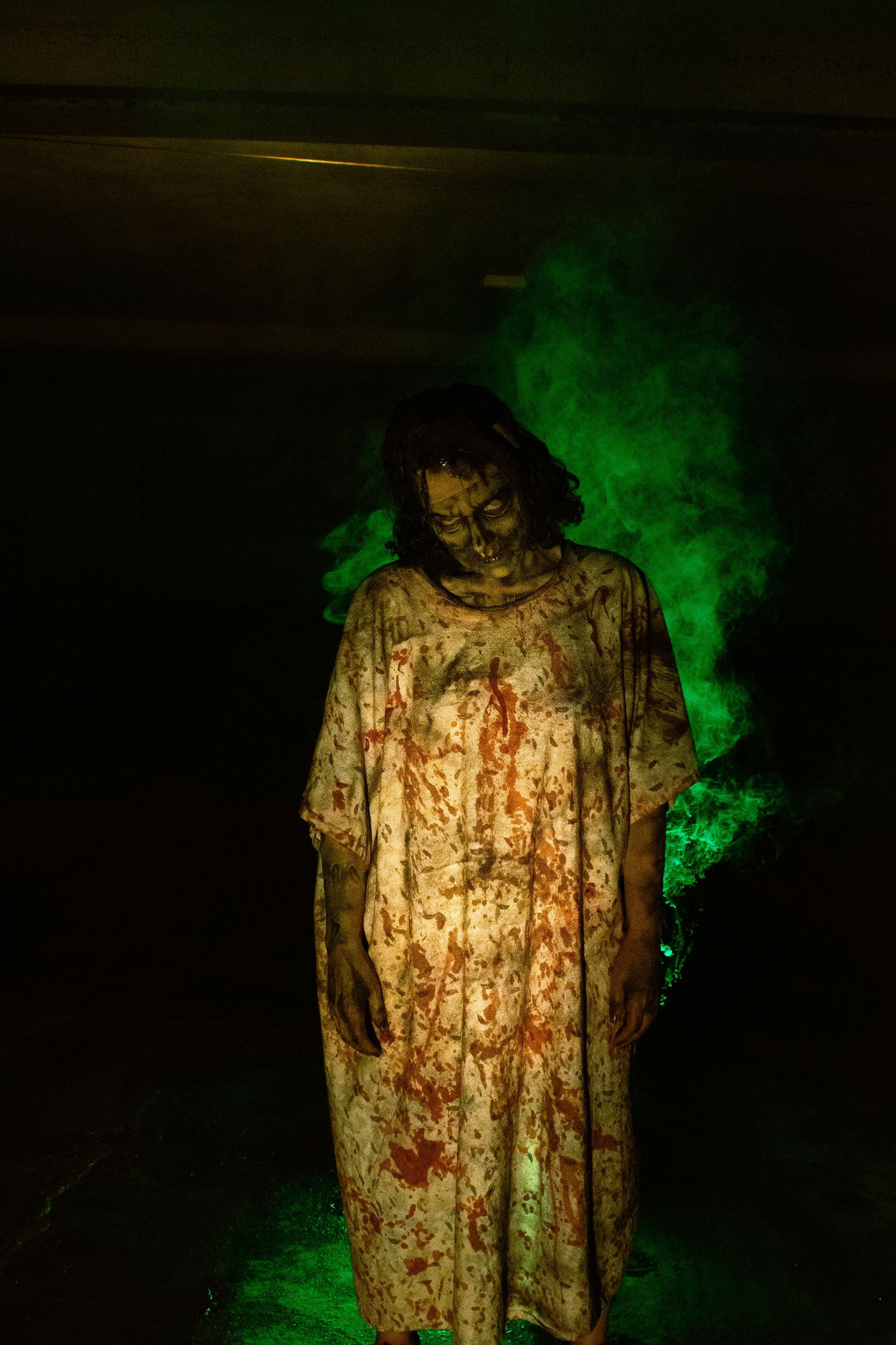 Bloody figure in a white gown standing in a dark room with green smoke behind it