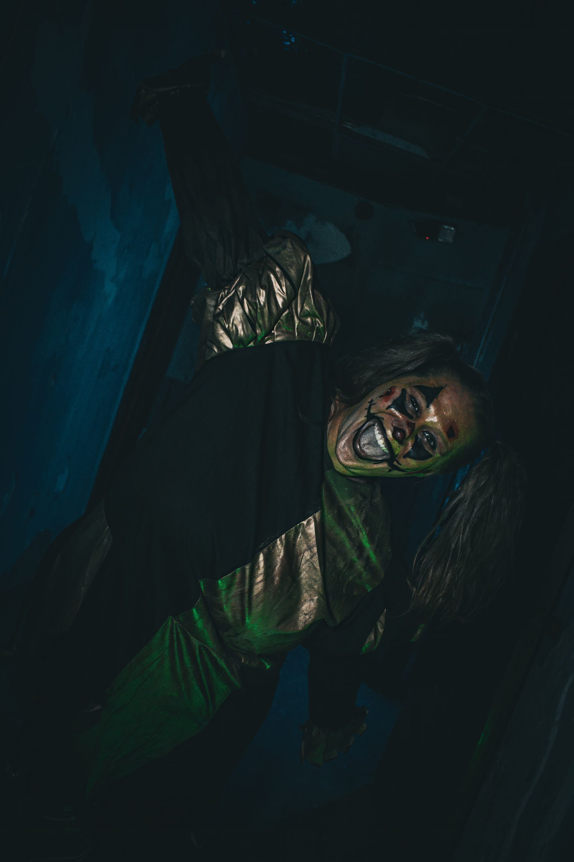 A grimacing person in a dark green outfit lies in shadowy blue light, with a tense, eerie expression.