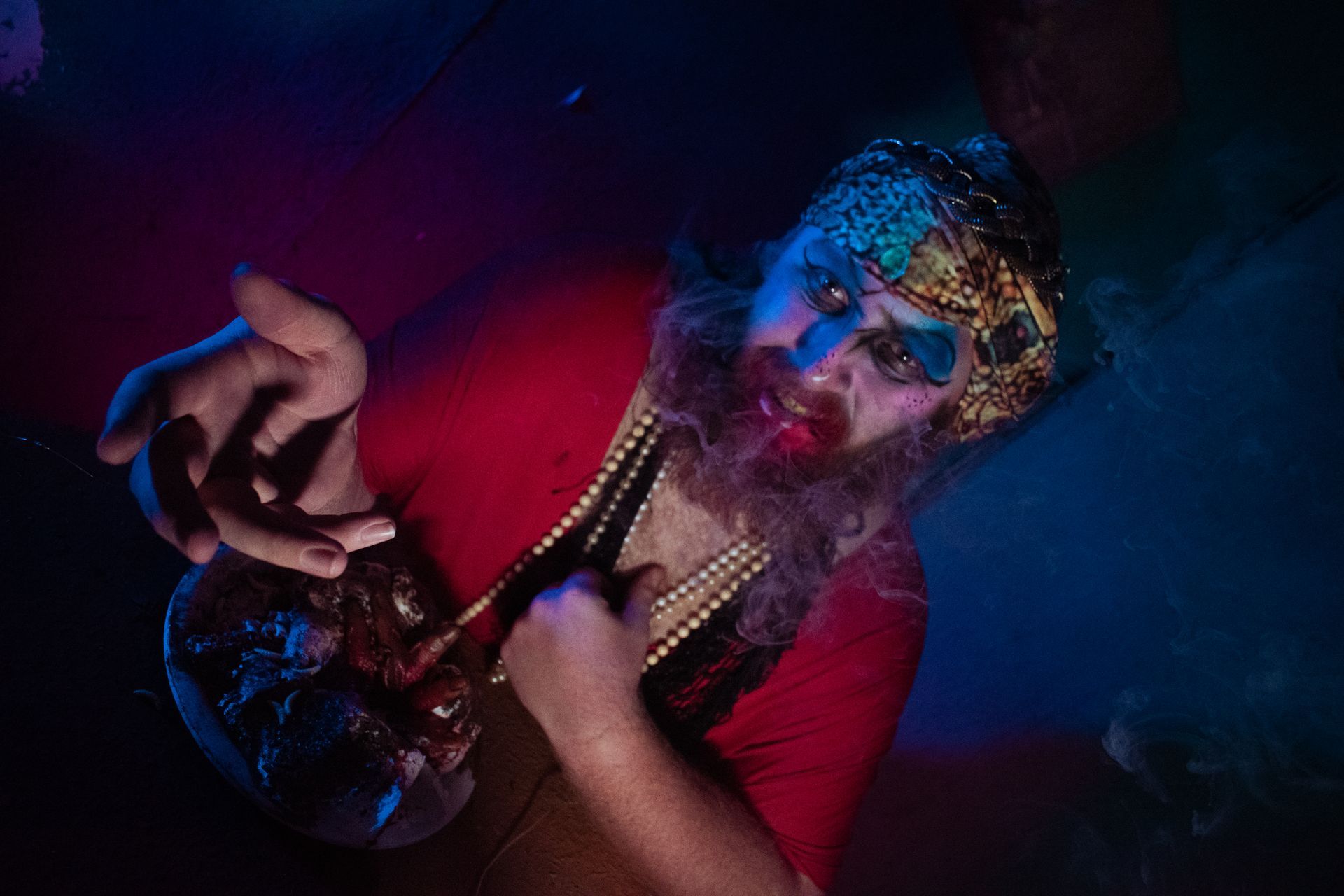 Person in red shirt and gold headpiece smoking under neon blue and red lighting