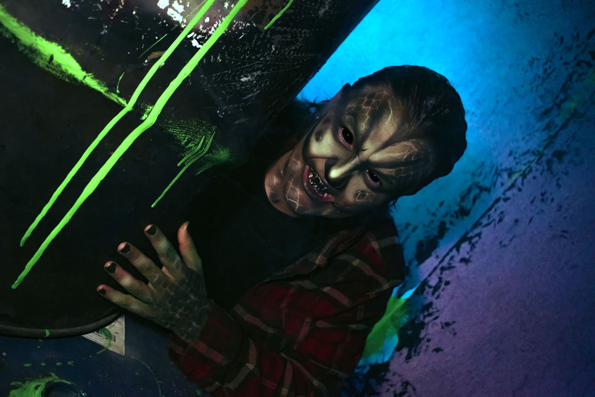 Person with face paint peeks from behind a large black object with neon green stripes in blue-purple lighting.