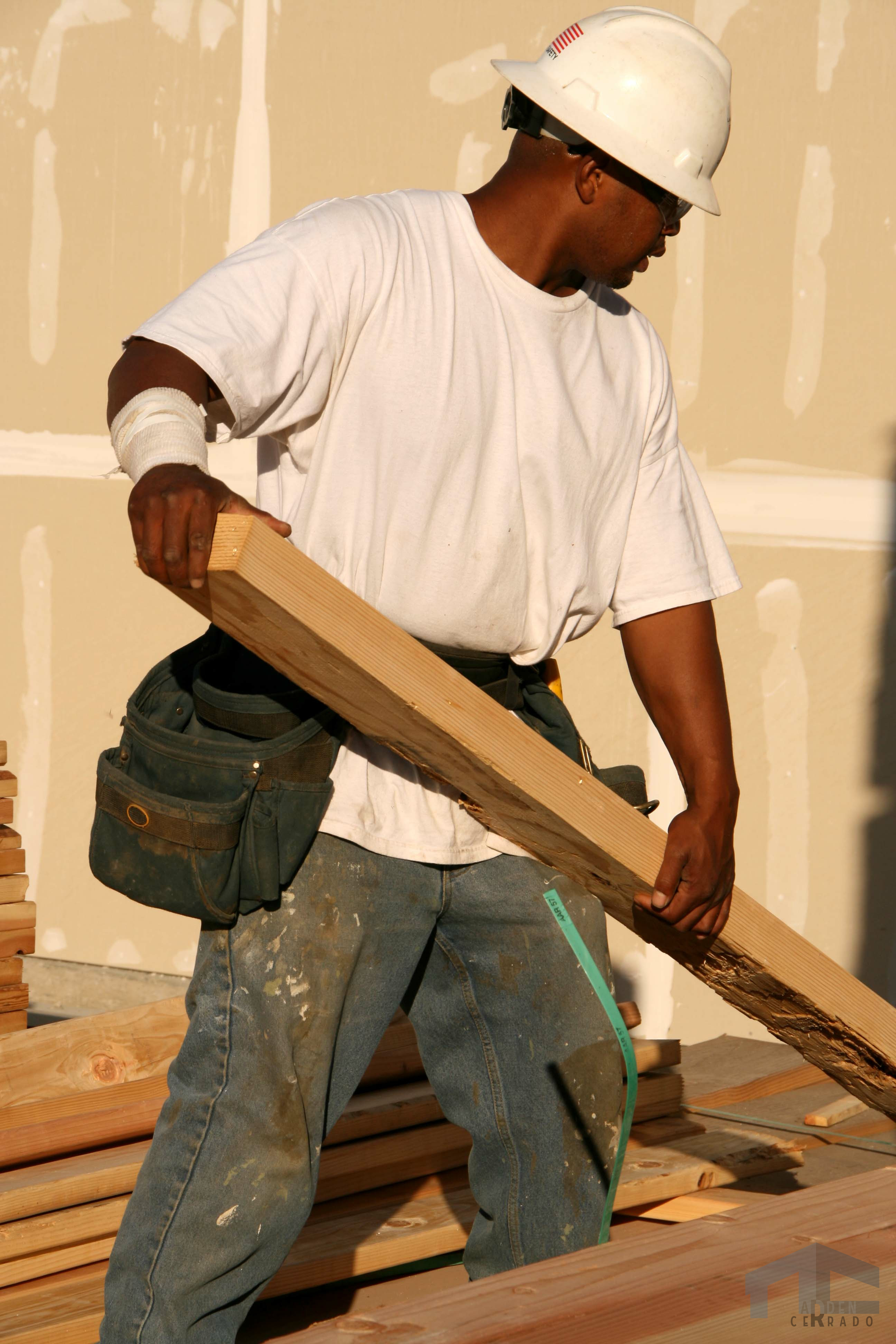 Construction worker carrying a wooden beam, wearing a white hard hat and tool belt.