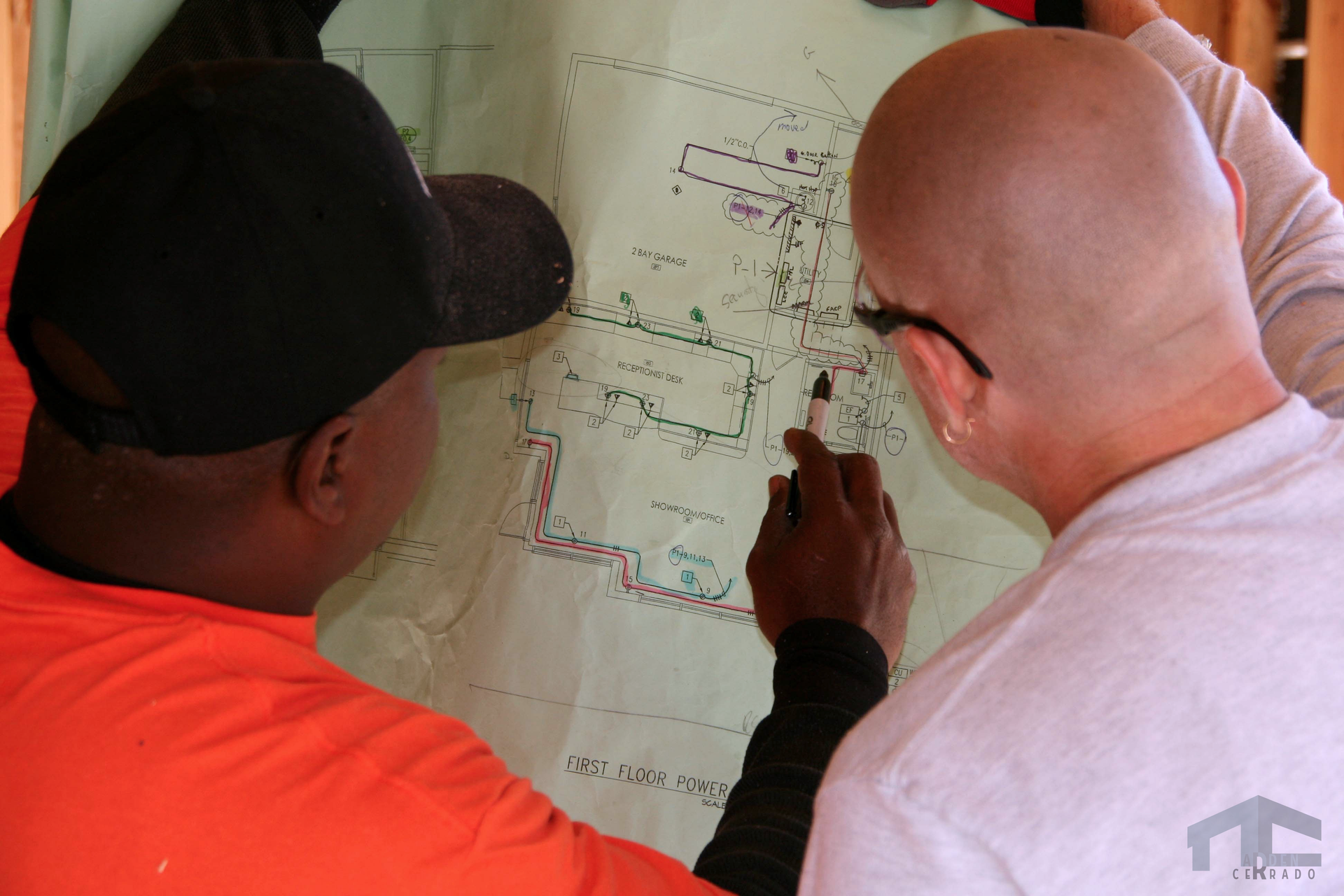 Two people examining a blueprint, one in an orange shirt and cap, the other with glasses, pointing with a marker.