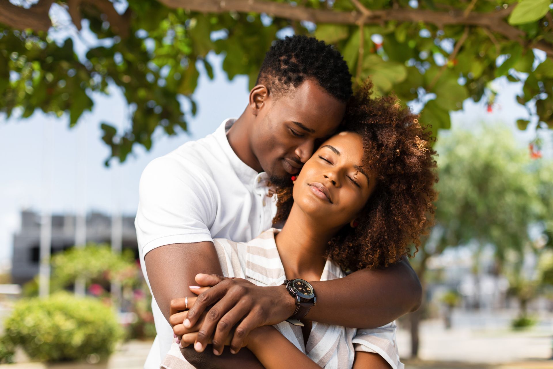 Couple embracing, man kissing woman's neck under a tree, outdoor setting.