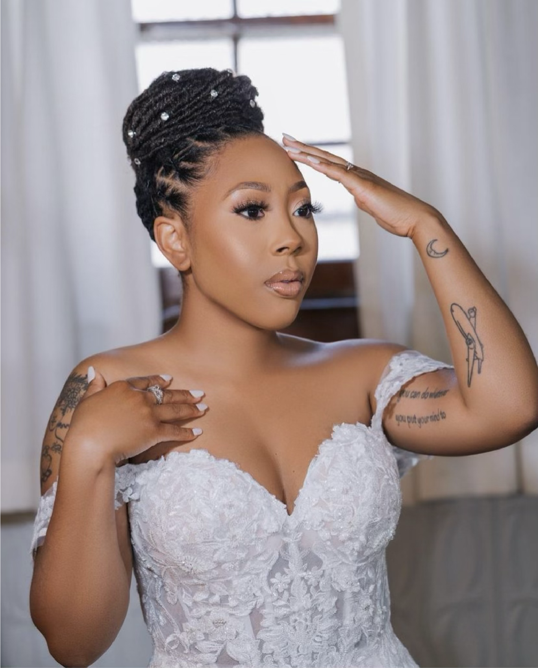 Woman in a white wedding dress, updo braided hair, hand on head, touching shoulder, tattoos visible.