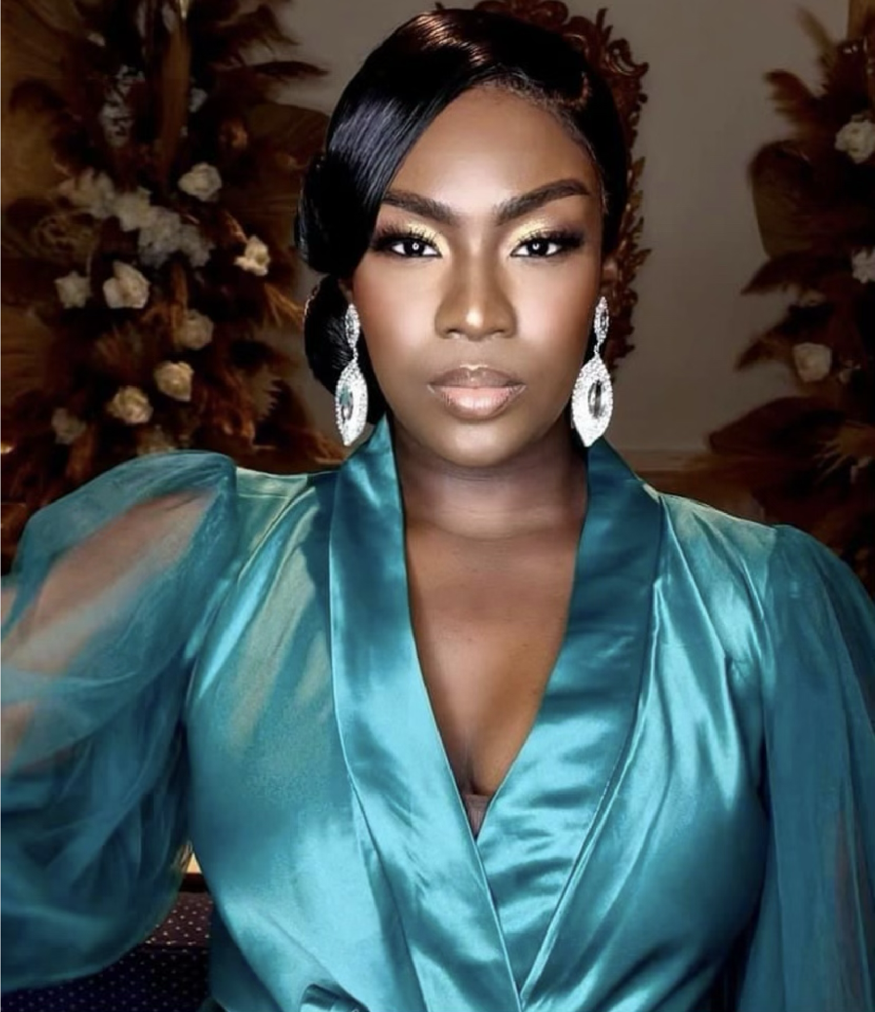 Woman in teal robe with diamond earrings, gold eye makeup, and black hair with a side part, facing the camera.