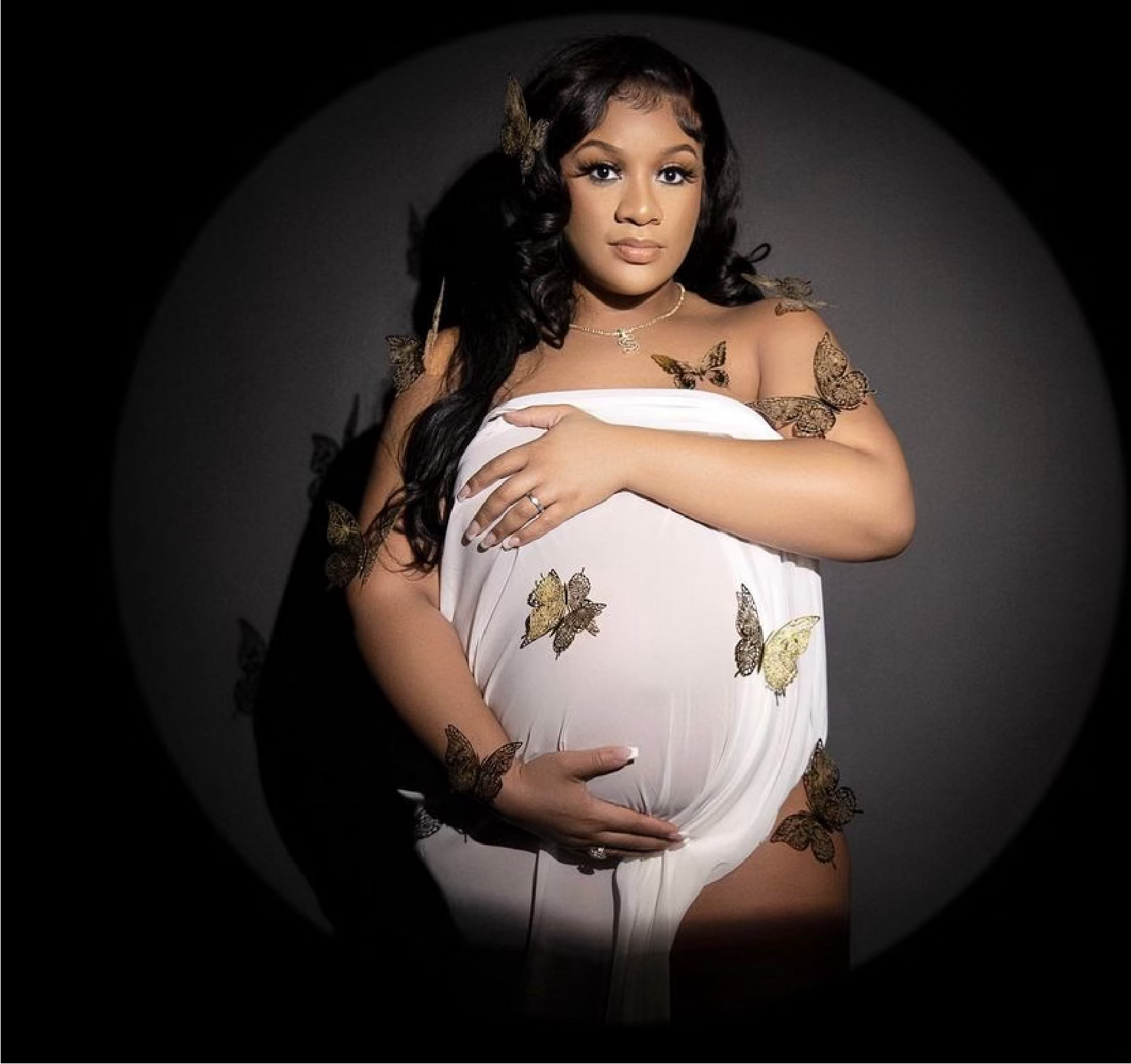 Pregnant woman in white drape, surrounded by butterfly decorations, hands on belly.
