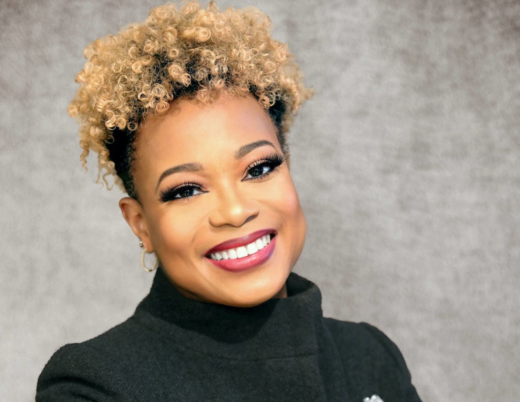 Woman with blonde curly hair smiles, wearing a black turtleneck.