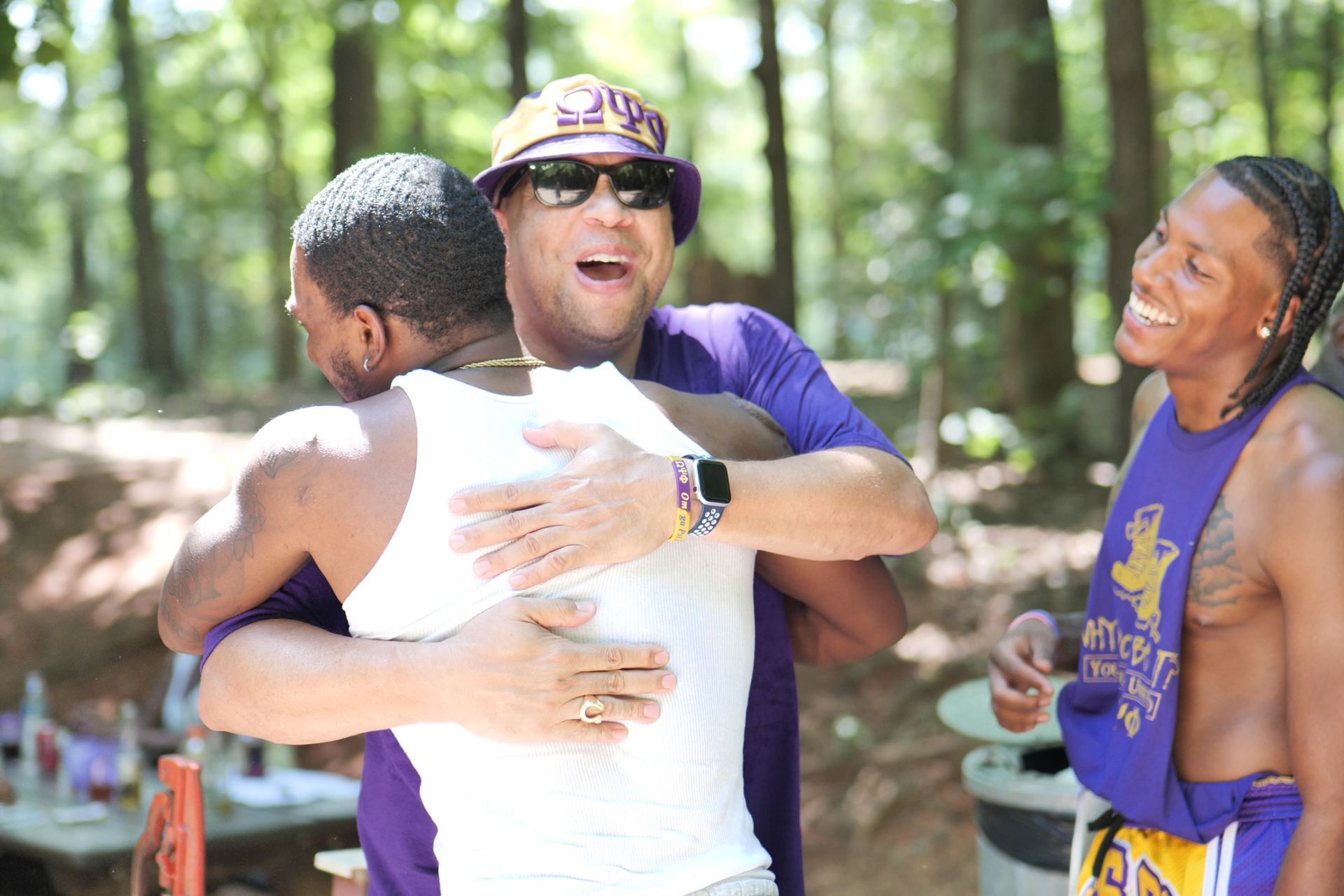 Three people hugging outdoors, wearing purple. One is in a white tank top, two are shirtless. Smiling faces, trees in background.