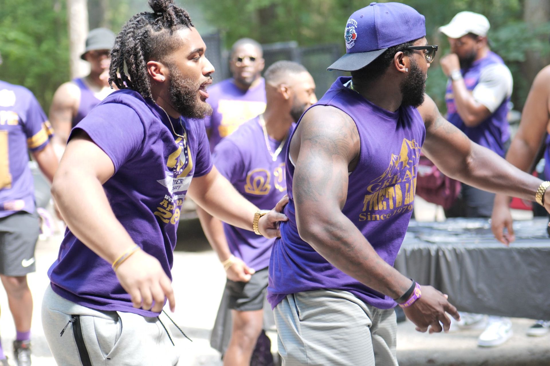 Two men in purple shirts dancing outdoors. Others in purple watch.