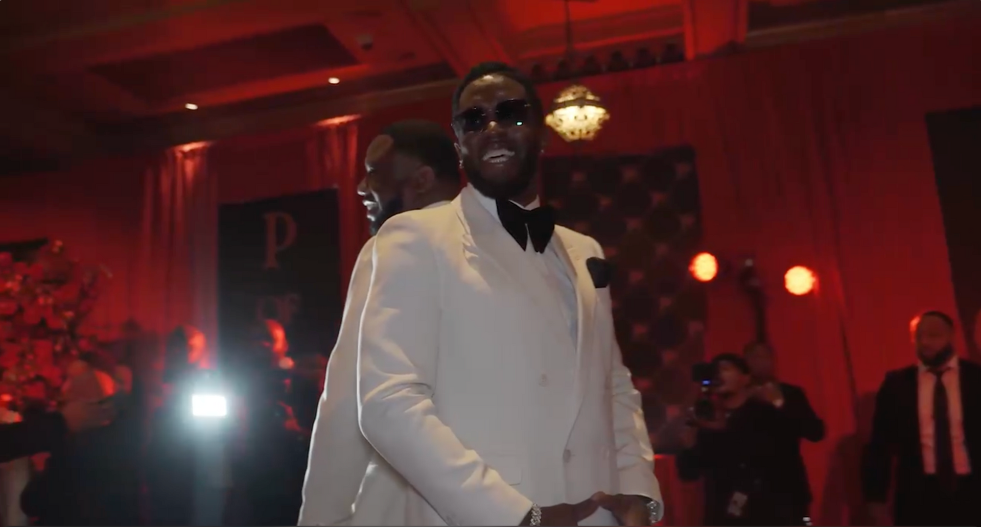 Man in a white tuxedo and sunglasses, standing in a room with red lighting, other people in the background.