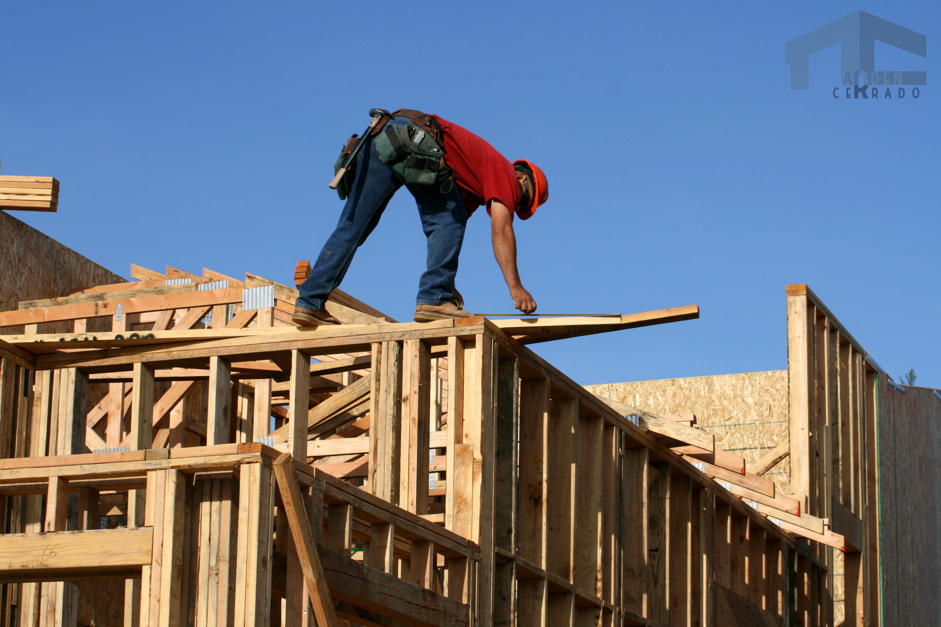 Construction worker on roof frame, measuring wood with a level, sunny blue sky.