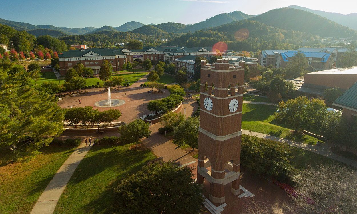 Brick clock tower on a campus with surrounding buildings, trees, and mountain backdrop under a bright sky.