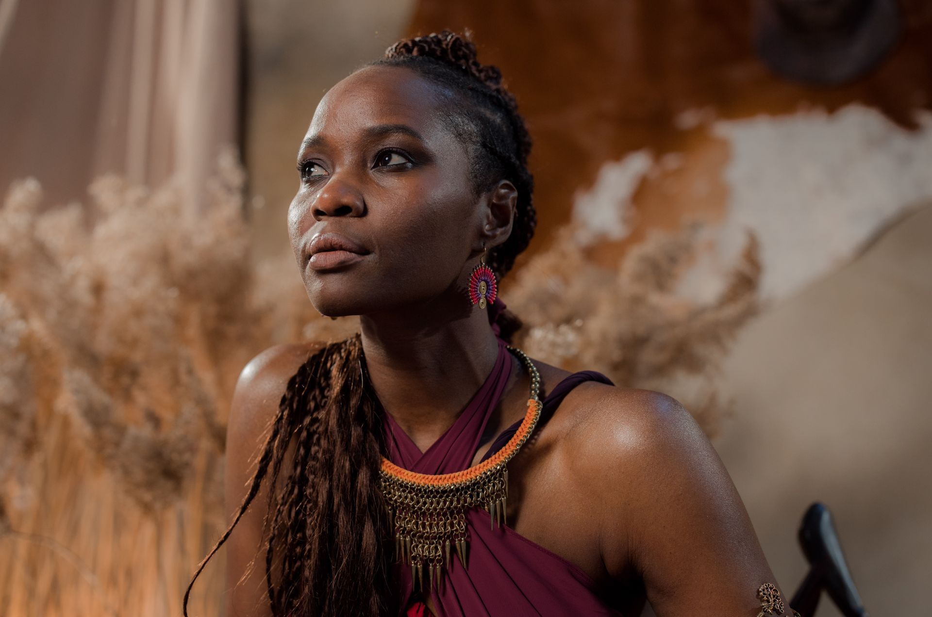 Woman with braided hair, wearing a burgundy dress and beaded necklace, looking off to the side.