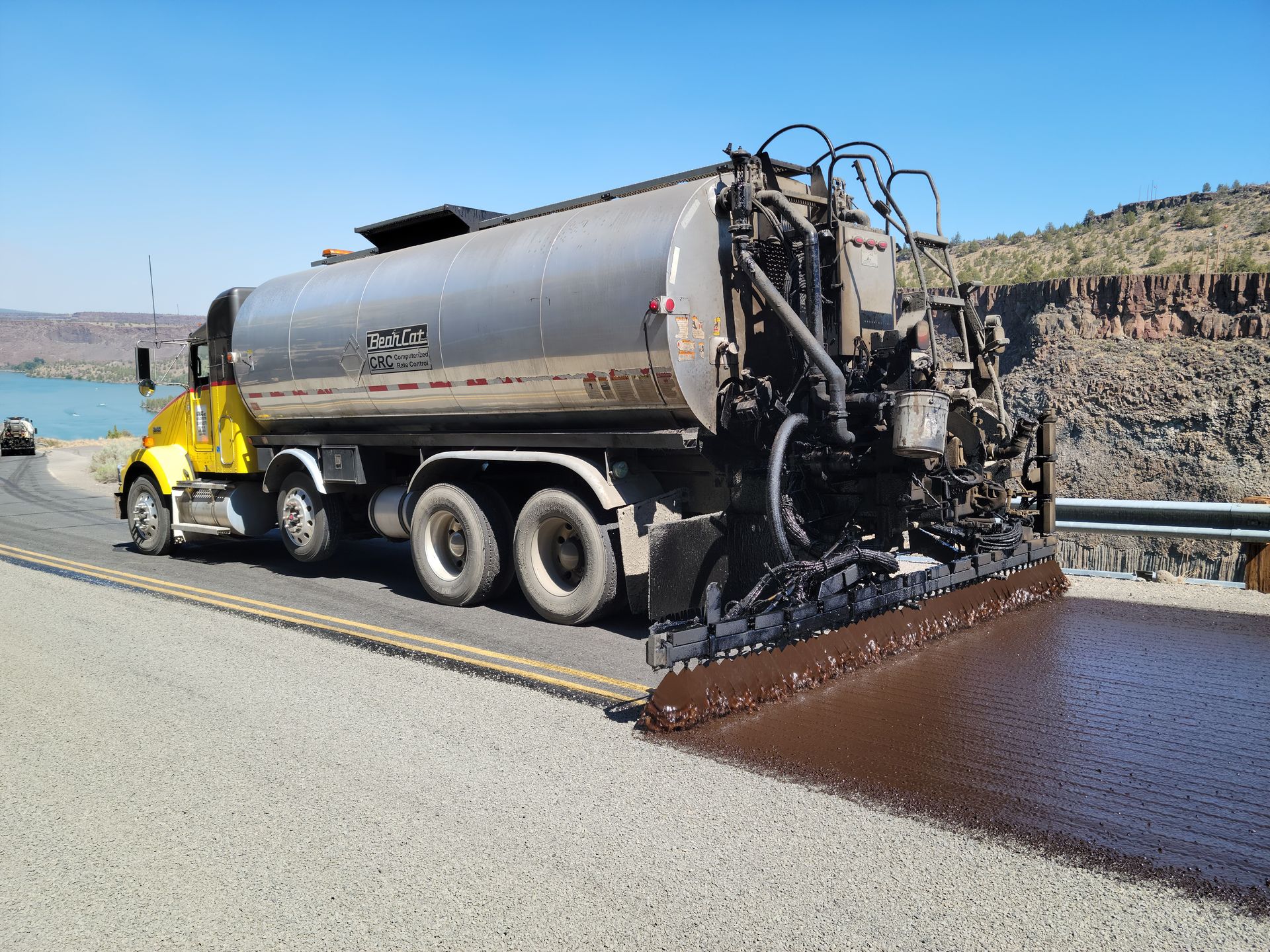 Yellow road truck spreading dark asphalt on a highway shoulder near rocky hills and water