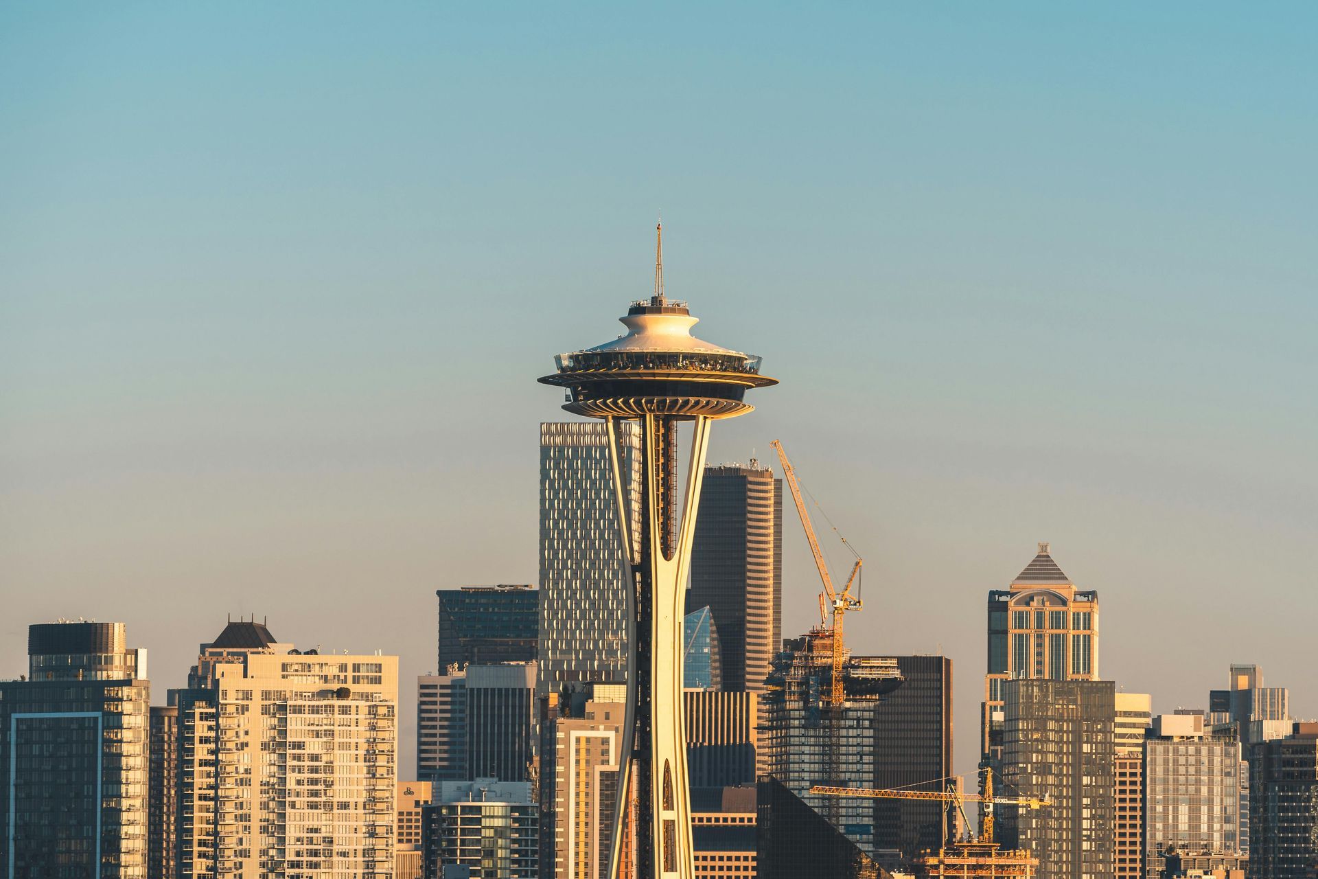 The Space Needle stands prominently in front of the Seattle skyline under a clear, pale blue sky at sunset.