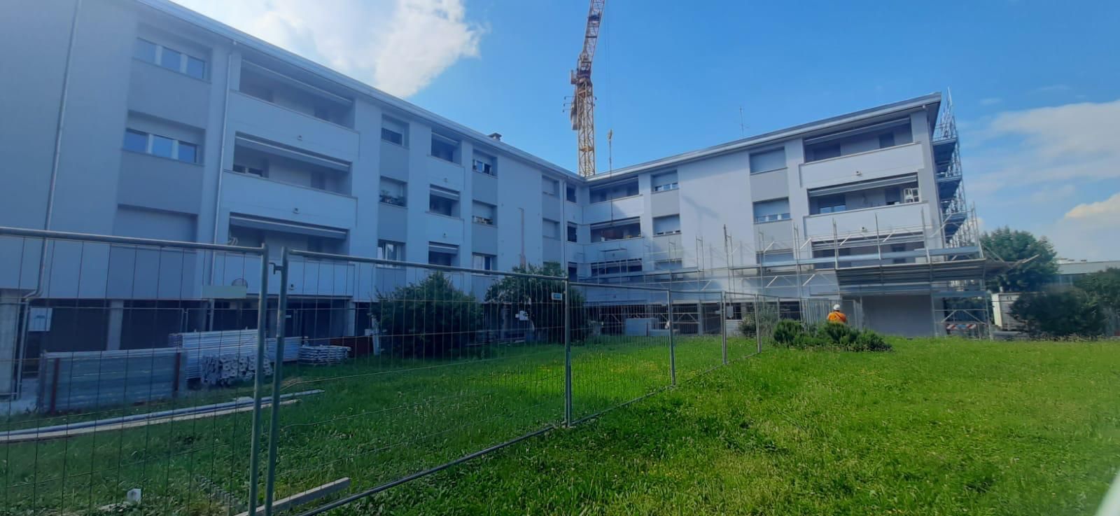 Palazzi residenziali bianchi accanto a un prato verde sotto un cielo azzurro, con una gru da cantiere sullo sfondo.