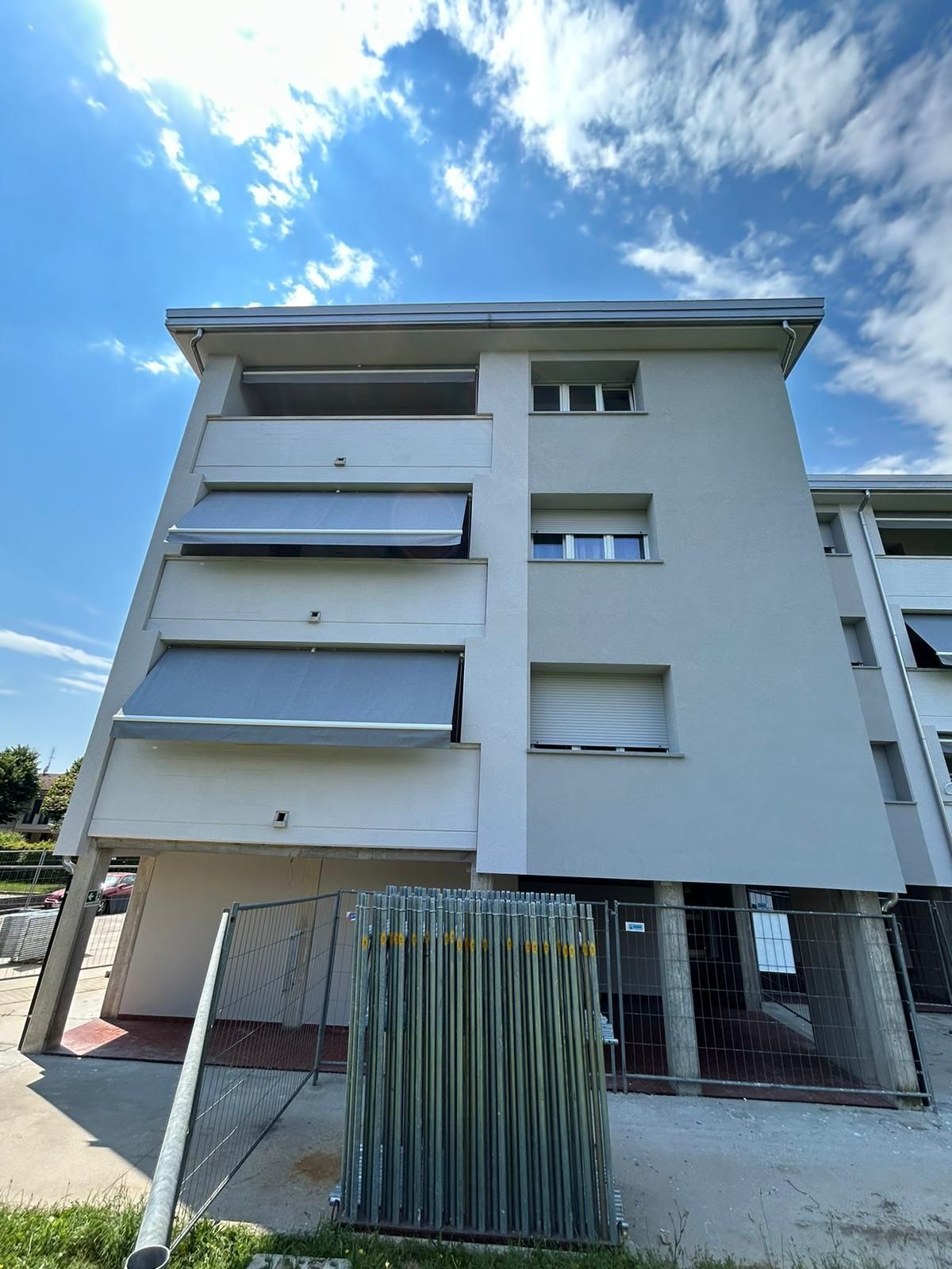 Moderno condominio con facciata bianca, balconi e ingresso recintato sotto un cielo azzurro.