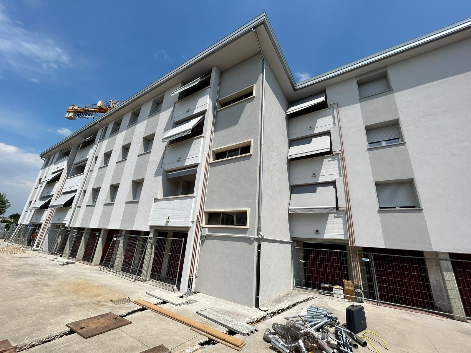 Moderno edificio residenziale in costruzione con facciata bianca e grigia, impalcature e piano terra aperto.
