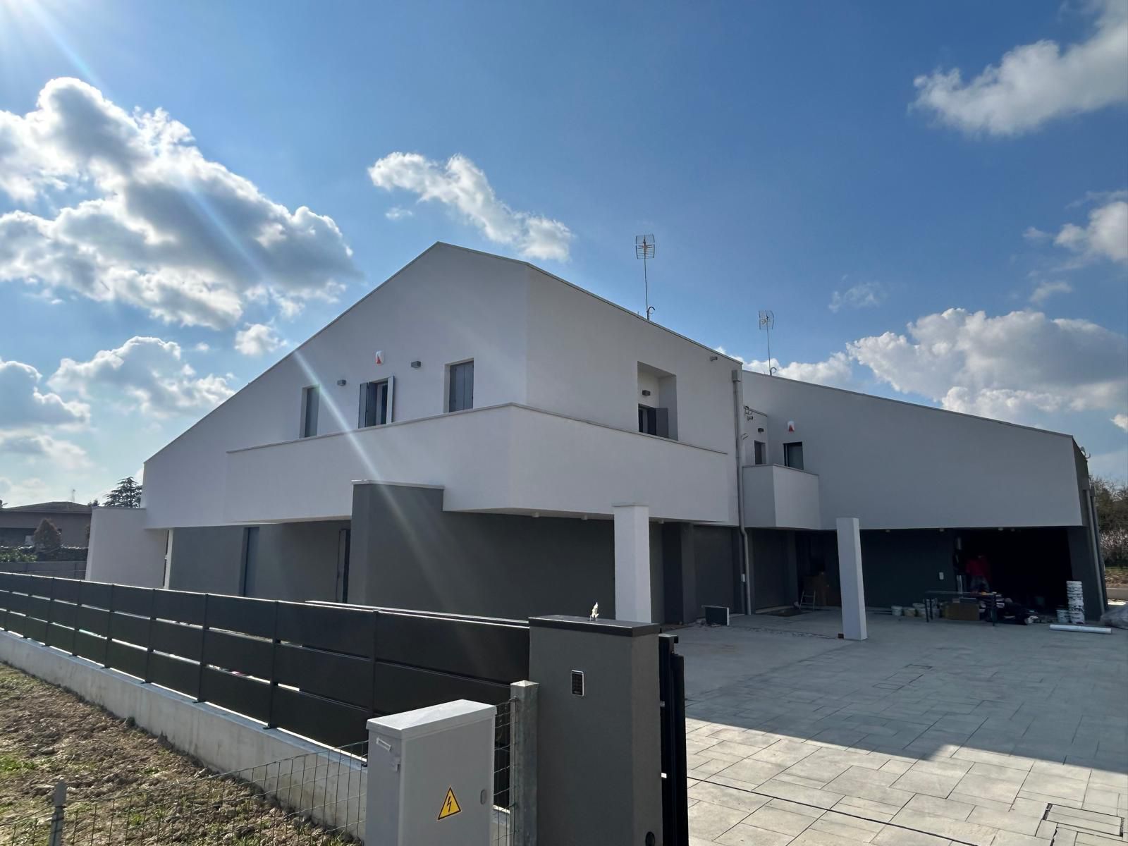 Casa moderna bianca a due piani con recinzione scura e vialetto pavimentato sotto un cielo parzialmente nuvoloso