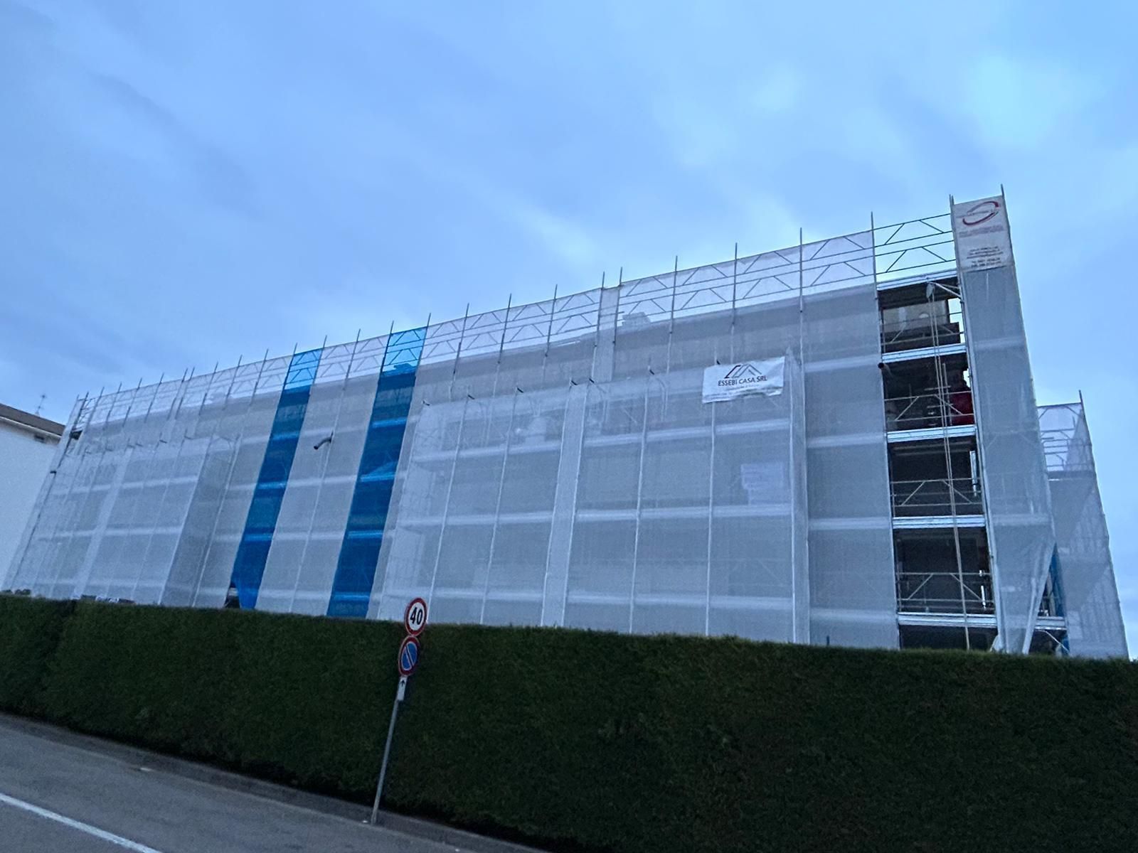 Grande edificio avvolto in una rete metallica bianca con strisce verticali blu, nascosto dietro una siepe sotto un cielo nuvoloso.