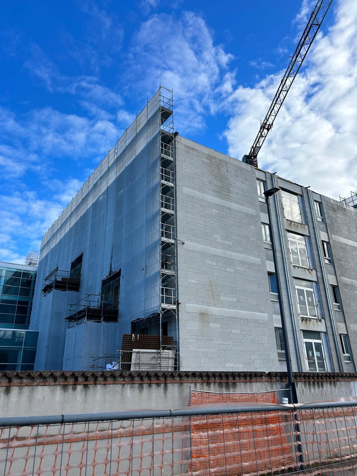 Esterno di un moderno edificio in costruzione, avvolto da una rete grigia, con una gru in volo sopra e il cielo azzurro sullo sfondo.