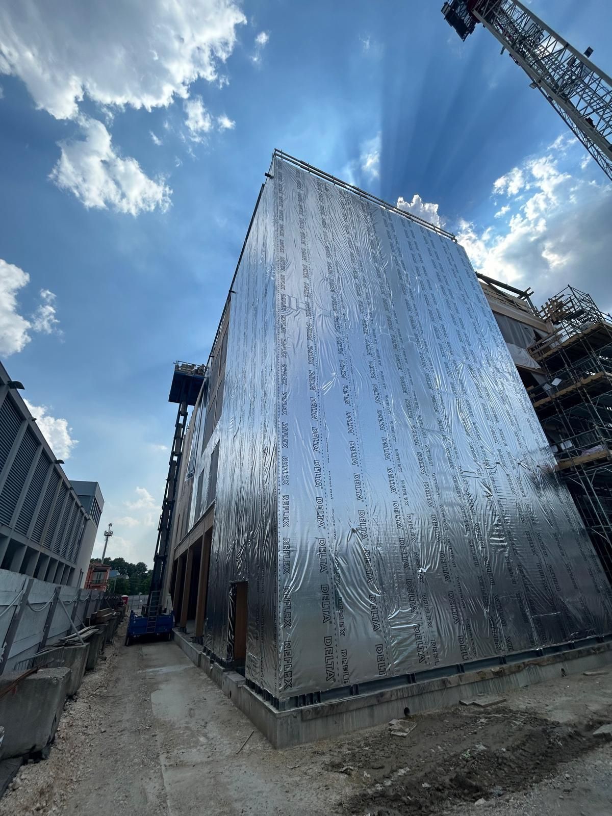 Un edificio lucido e specchiante in costruzione accanto a un sentiero sterrato e a una gru, sotto un cielo azzurro con nuvole.