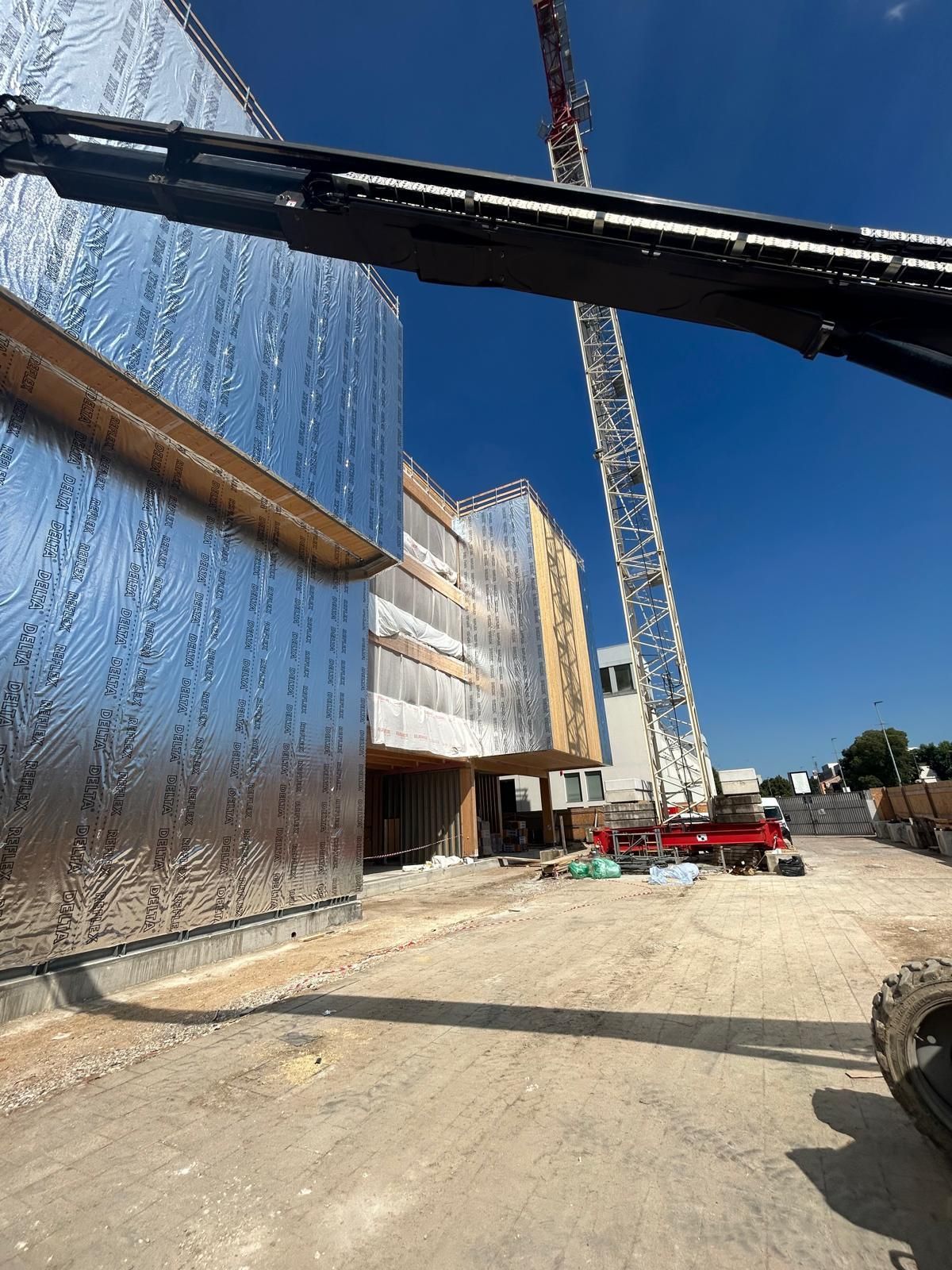 Cantiere edile con pannelli murali argentati riflettenti, gru e operai sotto un cielo azzurro limpido.